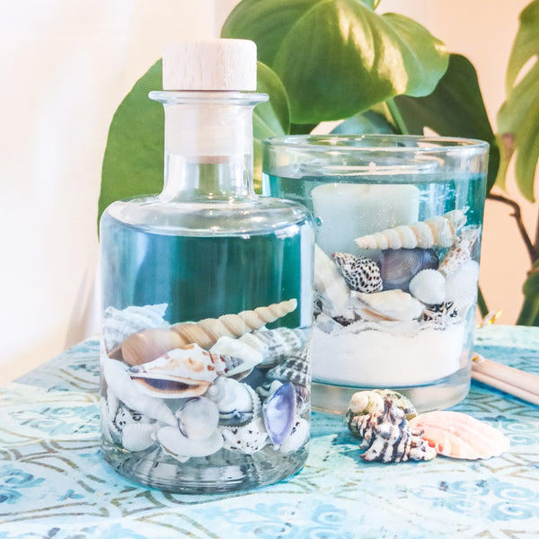 The blue sea reed diffuser is placed in front of the scented soy and gel  tumbler on a matching turquoise table top. Shells are scattered around and a large plant is in the background.