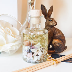 Coast reed diffuser. Apothecary style bottle filled with natural coloured shells and small white dried flowers is shown on a white dresser next to a white peony, a glass vase and bronze hare.