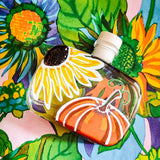 A clear glass bottle with a white cap, decorated with a large yellow sunflower and an orange pumpkin illustration, set against a colorful floral background.
