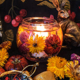 Autumn seasonal soy and gel wax fishbowl tumbler with vibrant dried flowers and mini pumpkins. The scented candle is lit, with a cosy glow against a dark floral background. Dried flowers and a bronze hare are placed next to the candle.