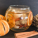 Festive orange and spices soy and gel wax fishbowl candle containing real dried orange slices, gold berries, cinnamon sticks and star anise. The scented candle is placed on a dark background next to dried oranges, cinnamon sticks and fir cones.