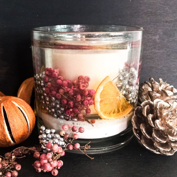 Sparkling cranberry soy and gel large tumbler. Scented container candle with pink and silver berries, dried orange slices on a base of snow white sand. The tumbler is placed on a dark background next to dried oranges, berries and pine cones.