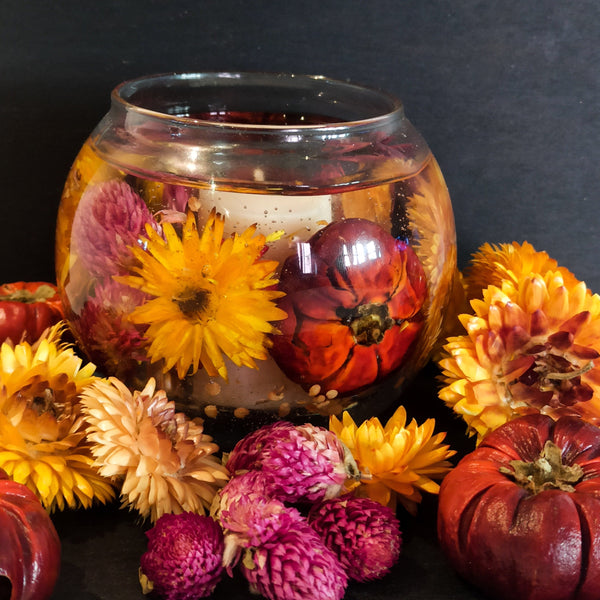 Autumn seasonal soy and gel wax fishbowl scented candle with vibrant dried flowers and mini pumpkins in vibrant autumnal hues is shown on a dark slate background surrounded by bright dried flowers and pumpkins.
