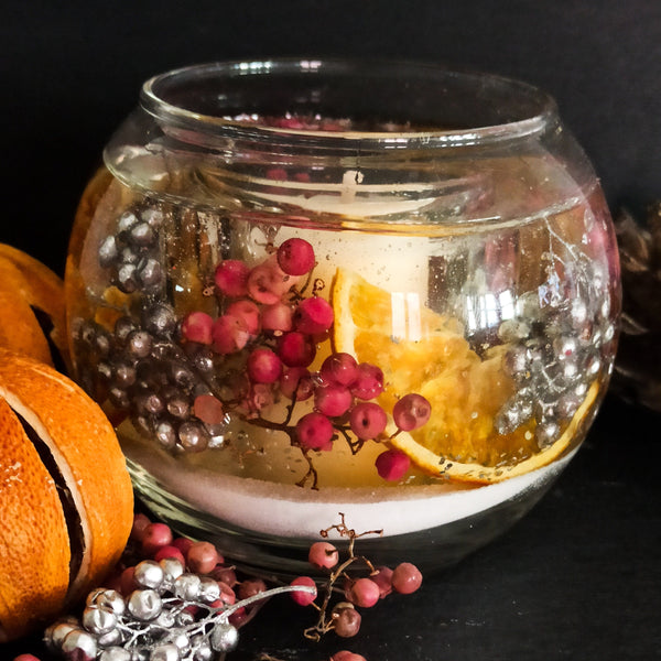 Sparkling cranberry soy and gel wax scented candle fishbowl design containing vibrant dried orange slices with pink and silver berries on snow white sand. The candle rests on a dark background next to oranges and berries.