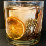 Festive orange and spices soy and gel wax tumbler, large scented candle with orange slices, gold berries, cinnamon sticks, star anise and crushed gold crystal on a dark slate background.