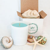 Turquoise fragranced large soy wax candle in a matte white candle jar on a white surface with a gift box and cotton bag surrounded by shells and a star fish.