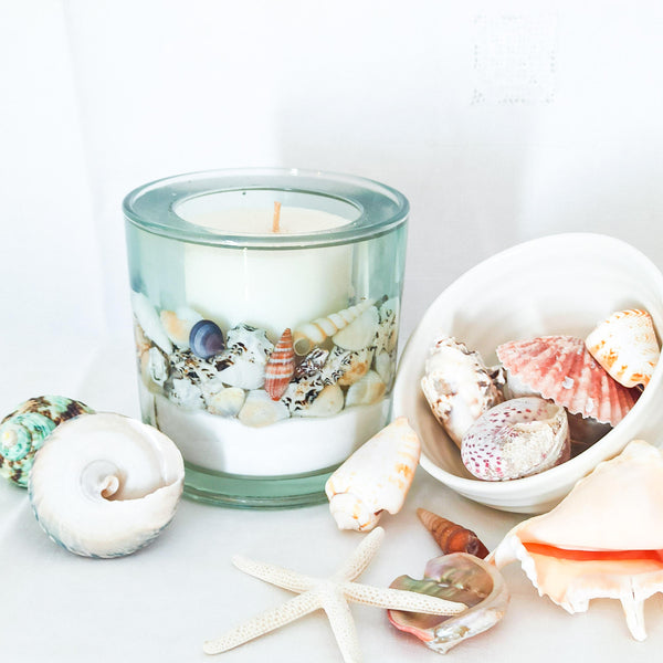 Large blue sea soy and gel wax scented ocean candle, handmade with real shells on a bed of pure white sand with a suspended in a transparent sea blue gel wax with a large white soy wax candle in the centre. The candle is shown on a white background next to a white dish filled with shells that are spilling out around the candle. The image is both fresh and relaxing reminiscent of tropical seas.