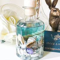 Blue sea ocean reed diffuser contains a mixture of small sea shells in an apothecary  shaped bottle with a turquoise blue coloured fragrance oil reminiscent of tropical seas off desert islands. The diffuser rests on a white surface near a bronze hare ornament and a hygge and hearth business card. A large white flower in a glass bowl and a glass vase are behind the diffuser giving a scene that is serene and relaxing whilst bringing thoughts of far flung holidays.