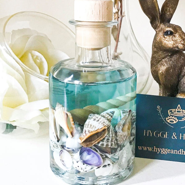 Blue sea ocean reed diffuser contains a mixture of small sea shells in an apothecary  shaped bottle with a turquoise blue coloured fragrance oil reminiscent of tropical seas off desert islands. The diffuser rests on a white surface near a bronze hare ornament and a hygge and hearth business card. A large white flower in a glass bowl and a glass vase are behind the diffuser giving a scene that is serene and relaxing whilst bringing thoughts of far flung holidays.
