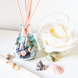 Teardrop shaped glass reed diffuser with natural coloured reeds placed in the bottle filled with blue fragrance oil and small seashells. Next to it is a round glass vase containing a large white rose, additional seashells are scattered on the white surface nearby, creating a soft, relaxing beach-inspired scene.