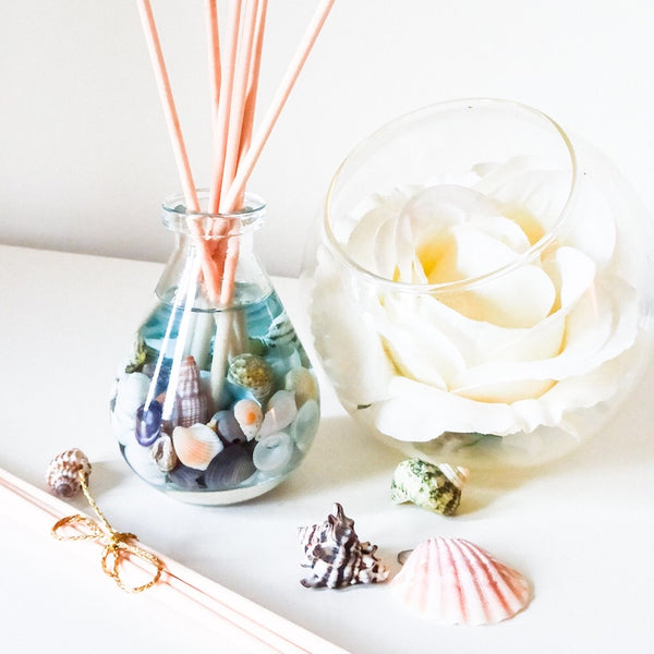 Teardrop shaped glass reed diffuser with natural coloured reeds placed in the bottle filled with blue fragrance oil and small seashells. Next to it is a round glass vase containing a large white rose, additional seashells are scattered on the white surface nearby, creating a soft, relaxing beach-inspired scene.