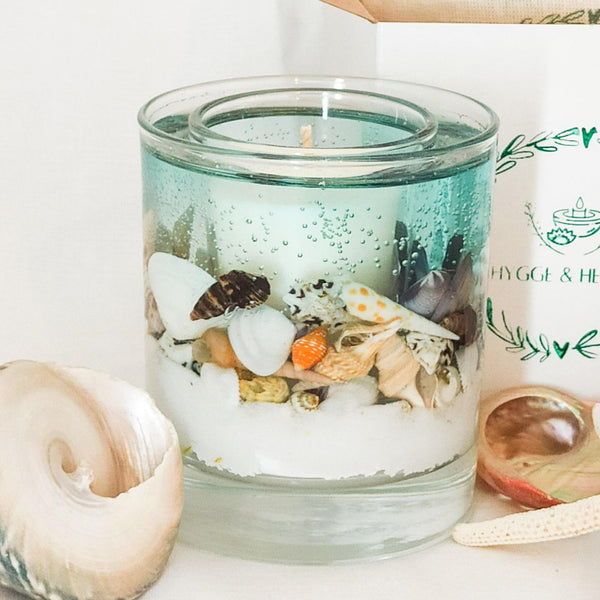 Blue Sea Ocean Candle, handmade soy and gel wax candle. Real shells are placed on a bed of white sand and encased in a sea blue soft gel wax with a white scented soy wax candle in the centre. The candle is on a white background in front of a gift box stamped with the Hygge and Hearth logo, while large shells are placed to the sides giving a tropical but relaxing feel to the scene.