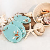 Blue Sea Ocean scented soy wax melts/room fresheners.  Three large oval shaped room fresheners in a shade of  aqua marine, decorated with small shells and a natural twine loop for hanging.  The room fresheners are on a white background resting on a brown  card giftbox surrounded by large shells, a white starfish and a bowl spilling large seashells, capturing the essence of blue seas and paradise islands.
