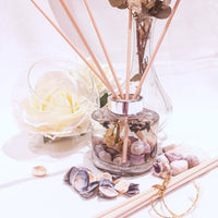 Lavender reed diffuser shown being used with natural coloured reed sticks. The circular glass diffuser contains clear fragrance oil containing small seashells in hues of purple, small dried white flowers and sprigs of dried lavender. A large white rose in a clear bowl is next to the diffuser with a taller glass vase, purple seashells are scattered on the white surface near to more diffuser reeds, to set a scene of natural elegance and relaxation.