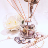 Lavender reed diffuser shown being used with natural coloured reed sticks. The circular glass diffuser contains clear fragrance oil containing small seashells in hues of purple, small dried white flowers and sprigs of dried lavender. A large white rose in a clear bowl is next to the diffuser with a taller glass vase, purple seashells are scattered on the white surface near to more diffuser reeds, to set a scene of natural elegance and relaxation.