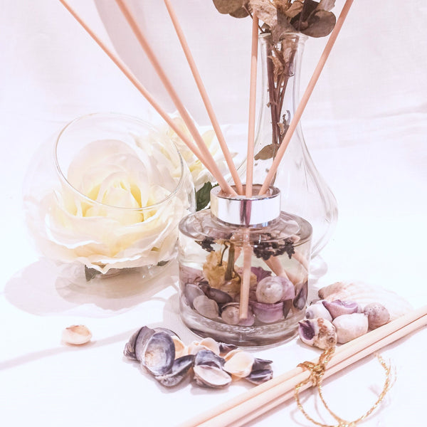 Lavender reed diffuser shown being used with natural coloured reed sticks. The circular glass diffuser contains clear fragrance oil containing small seashells in hues of purple, small dried white flowers and sprigs of dried lavender. A large white rose in a clear bowl is next to the diffuser with a taller glass vase, purple seashells are scattered on the white surface near to more diffuser reeds, to set a scene of natural elegance and relaxation.