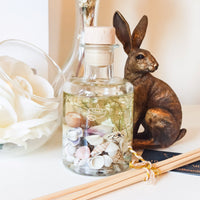 A  white dressing table has a large white flower, glass vases and a bronze hare ornament. At the centre the Coast 200ml diffuser is shown in its apothecary  bottle filled with seashells and small white dried flowers, creating a relaxing, nature inspired decorative scene. 