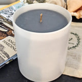 Coast handmade scented candle. Made from dark grey soy wax the large 30cl candle is housed in an elegant matte white candle jar. The candle is on a slate surface with a book and large seashells in the background.