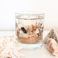 Coast scented gel and soy wax candle crafted with natural sand and shells encapsulated in a soft gel with a soy votive core. The candle is placed on a white table with soft coloured large shells surrounding it.