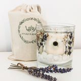 Reusable botanical candle with shells, lavender and daisies resting on a white surface with dried lavender and a printed white cotton drawstring bag.
