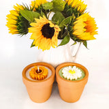 Two terracotta pots containing soy wax candles, one with a yellow sunflower motif and one with a white daisy motif in a reusable terracotta flower pot. The handmade candles are placed in front of a white vase containing sunflowers.