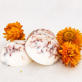 Three large white handcrafted botanical soy wax melts with coloured petal embeds are on  a white surface next to three deep yellow dried flowers