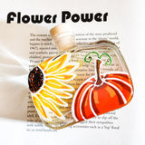 A clear glass bottle with a cork stopper, decorated with a painted flower and leaf motif in yellow, orange, red, green, and brown, with the words 'Flower Power' printed above on a light background overprinted with book text.