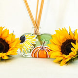 Large refillable handcrafted reed diffuser with square bottle, hand painted  with a sunflower and pumpkin and containing bright green fragrance oil. The diffuser is on a white background with two large sunflowers on each side