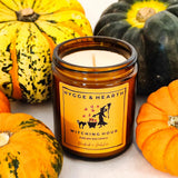 Hygge and Hearth Witching Hour pure soy wax candle in an amber candle jar with an orange label with a silhouette of a witch and cat illustration. The candle is on a white background surrounded by four squashes in various colours.