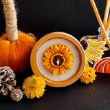 Hygge and Hearth soy wax Sunflower Candle in a terracotta plant pot.  The candle is placed on its side with the yellow sunflower wax motif facing forward on a dark background surrounded by a handpainted reed diffuser, dried flowers, pine cones and a bright orange textile pumpkin.