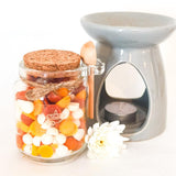 Jar of mini wax melts in orange, yellow and white with a cork lid, wooden spoon, and grey wax melt burner on a white background.