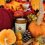 An amber jar candle labeled 'Hygge & Hearth Spiced Pumpkin' surrounded by autumn decorations including fabric pumpkins, autumn leaves, pinecones, and cinnamon sticks.