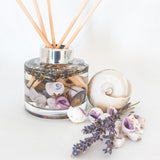 Lavender botanical reed diffuser with purple toned shells and real sprigs of lavender for a hygge home. The handcrafted diffuser is on a white background next to small purple and white shells, a large white shell and lavender stems giving a relaxing but fresh  ambience.