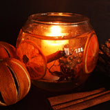 Round botanical candle bowl with orange slices cinnamon sticks and gold berries encapsulated in a clear gel wax with a soy wax votive burning at the centre. The handmade statement candle gives a cosy amber glow against a black background. The candle is surrounded by dried oranges and cinnamon sticks.