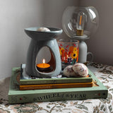 A peaceful and relaxing scene of a nightstand covered in an antique table cloth in muted cottagecore tones. Three books are on the nightstand with a soy wax burner which is melting a soy wax melt. A rustic handpainted floral candle holder and an old fashioned lamp are also on the table giving a rustic, hygge ambience. 