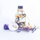 Botanical lavender fragranced reed diffuser with shells and dried flowers in an apothecary style bottle, on a white surface surrounded by dried lavender, a large shell and purple semi-precious stones.