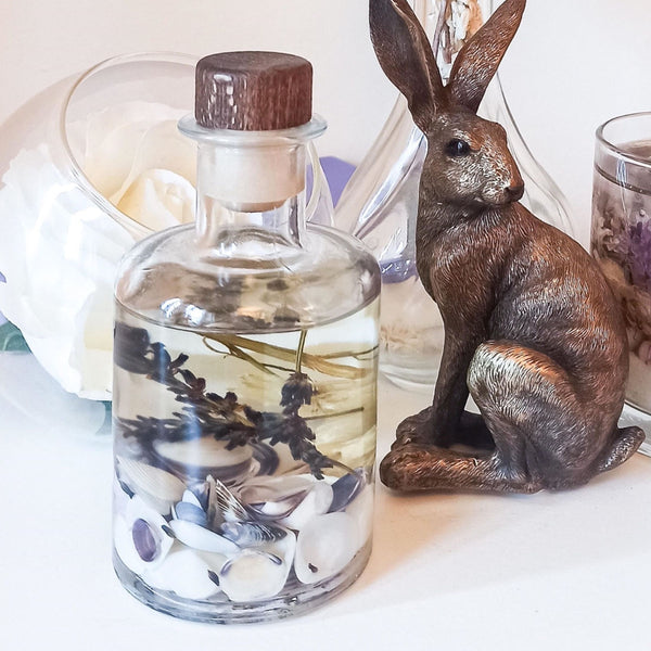 Lavender fragranced 200ml  reed diffuser in a clear apothecary style bottle with purple shells, dried lavender and white flowers suspended in the scented oil. The diffuser bottle is placed on a white surface next to a bronze hare ornament, with a glass bowl with a large white flower, a glass vase and a lavender botanical soy and gel wax candle ust in view, which gives a fresh but relaxing scene.