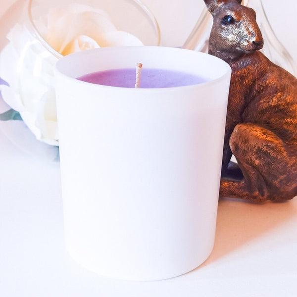 Lavender scented soy wax candle in a stylish, minimalist white container which compliments the lavender coloured candle. The candle is displayed on a white surface next to a decorative bronze hare and a large white rose in a glass bowl, creating a cosy, relaxing but elegant setting.