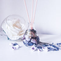 Reed diffuser with natural coloured reeds placed in the glass teardrop shaped bottle, which contains fragrance oil, seashells in shades of purple, dried white flowers and stems of lavender. Next to the diffuser is a bowl with a large white rose, purple shells and lavender stems are strewn nearby on the white surface creating a lavender hued, fresh, relaxing scene.