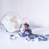 Reed diffuser with natural coloured reeds placed in the glass teardrop shaped bottle, which contains fragrance oil, seashells in shades of purple, dried white flowers and stems of lavender. Next to the diffuser is a bowl with a large white rose, purple shells and lavender stems are strewn nearby on the white surface creating a lavender hued, fresh, relaxing scene.