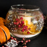 Festive refillable botanical candle containing pink and silver berries and dried orange slices encapsulated in a clear gel wax with a burnable soy wax candle core in a bowl shaped candle glass. The candle stands out on a dark background next to dried oranges and pink and silver berries. 