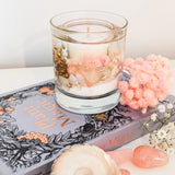 Pink Champagne soy and gel wax container candle decorated with real white shells, pastel pink and white flowers and gold berries is placed on a book with a grey, black and gold cover. A large shell. pink and white dried flowers and rose quartz are scattered around.