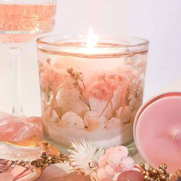 Pink Champagne luxury soy and gel wax candle glows casting a soft pink light. The candle contains white shells, pink, white and gold flowers, encapsulated in a gel wax with a soy candle core. The large handmade statement candle is placed between the Pink Champagne soy wax candle and a decorative glass of pink champagne with dried pastel coloured flowers and rose quartz scattered around. 