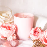 Pink champagne scented soy wax candle handmade using pastel pink wax in a pastel pink cylindrical candle jar. The candle is on a white table surrounded by white and pale pink flowers and a sprinkling of gold berries, creating a soft pink, romantic theme.