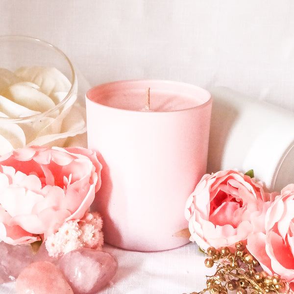 Pink champagne scented soy wax candle handmade using pastel pink wax in a pastel pink cylindrical candle jar. The candle is on a white table surrounded by white and pale pink flowers and a sprinkling of gold berries, creating a soft pink, romantic theme.