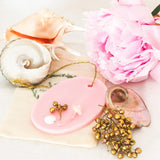 Pastel pink soy wax melt decorated with gold and white botanicals is on a white background surrounded by a large pink peony, gold berries and large sea shells. 