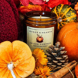 A Hygge & Hearth 'Spiced Pumpkin' soy candle in an amber glass jar, surrounded by autumn-themed props including miniature pumpkins, a pinecone, cinnamon sticks, and orange flowers.
