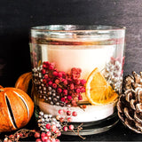 Large botanical festive sparkle botanical candle with pink and silver berries and orange slices enveloped in a soft gel wax with a large burnable soy wax candle in the centre. The candle is placed on a dark background surrounded by pine cones, dried oranges and colourful berries.