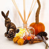 A clear glass reed diffuser bottle with a wrapped reed stem, decorated with a sunflower and orange pumpkin illustration. The diffuser is styled with autumn props including a bronze rabbit figurine, pinecones, a small orange pumpkin, yellow flowers, cinnamon sticks, and lavender on a white background.