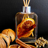 Winter Glow botanical reed diffuser with dried orange slices and a cinnamon stick immersed in fragrance oil and housed in a sleek tall rectangular bottle with rounded corners and  a silver collar.
The vibrant diffuser is striking against a black background and is surrounded by dried oranges, cinnamon sticks and gold berries.
